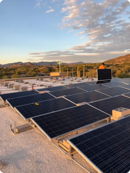 Rooftop solar panels on a flat roof with a desert landscape and mountains in the background during sunset. A worker stands near equipment on the roof.