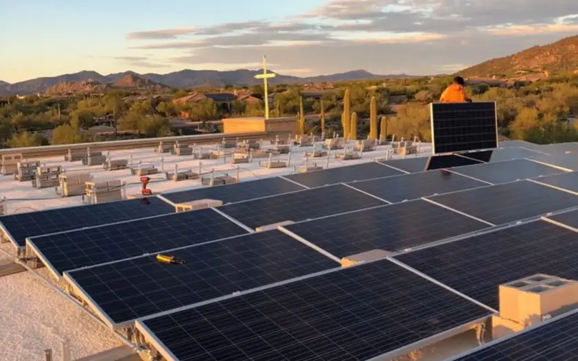 Rooftop solar panels on a flat roof with a desert landscape and mountains in the background during sunset. A worker stands near equipment on the roof.
